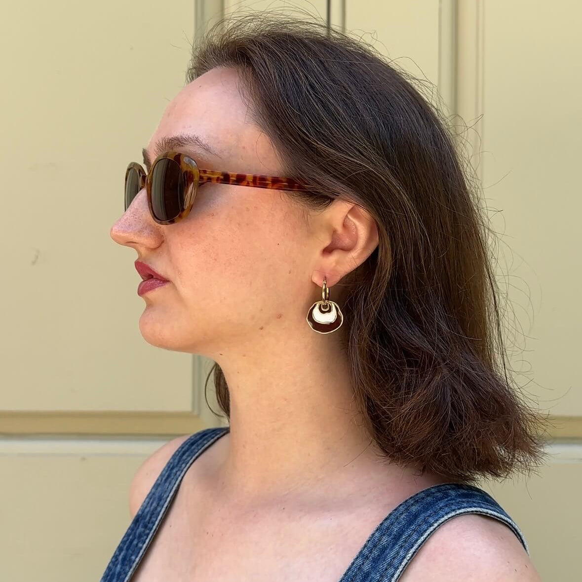 A woman in profile wearing tortoiseshell sunglasses with a gold-toned organic shaped circular drop earring, dressed in blue denim overalls against a beige paneled wall. The earring has brown and white enamel.
