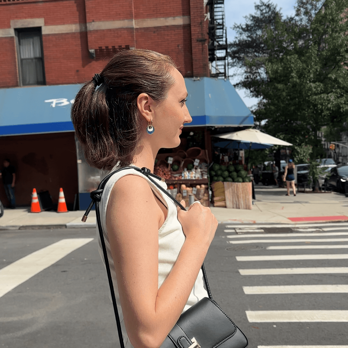 A woman stands on a city crosswalk wearing a structured black leather shoulder bag with a gold-tone buckle, paired with a white sleeveless top and a dark pleated skirt. Brick buildings, trees, and street elements are visible in the background. She is wearing a gold hoop earring with a white and blue concentric enamel dangle design