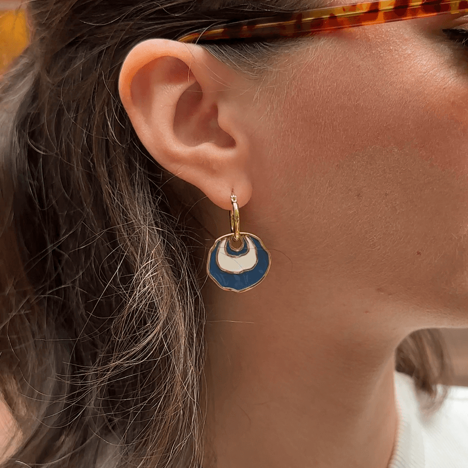 A woman in profile wearing tortoiseshell sunglasses and a white ribbed henley top, she is wearing a gold hoop earring with a white and blue concentric enamel dangle design