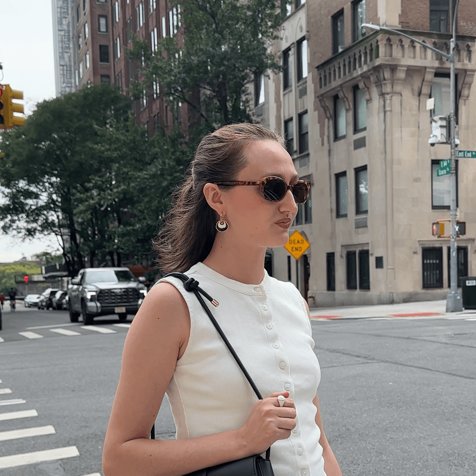 A woman wearing a white sleeveless top, a dark pleated mini skirt, and sunglasses walks across a city street crosswalk, carrying a black shoulder bag. She is wearing a gold hoop earring with a white and black concentric enamel dangle design.