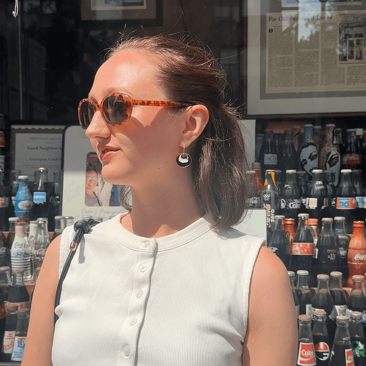 A woman wearing a sleeveless white top and tortoiseshell sunglasses stands in front of a shop window displaying vintage bottles. She is wearing a gold hoop earring with a white and black concentric enamel dangle design.