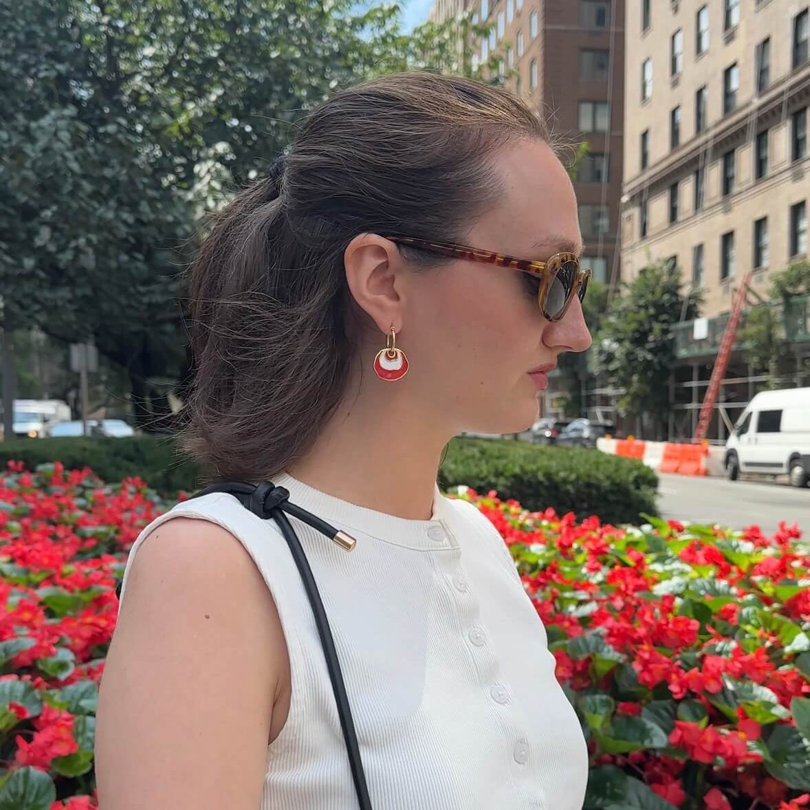A woman in a white sleeveless top and sunglasses stands outdoors in front of a flower bed with red flowers and a city building. She is wearing a gold hoop earring with a white and red concentric dangle design and carrying a black shoulder bag.