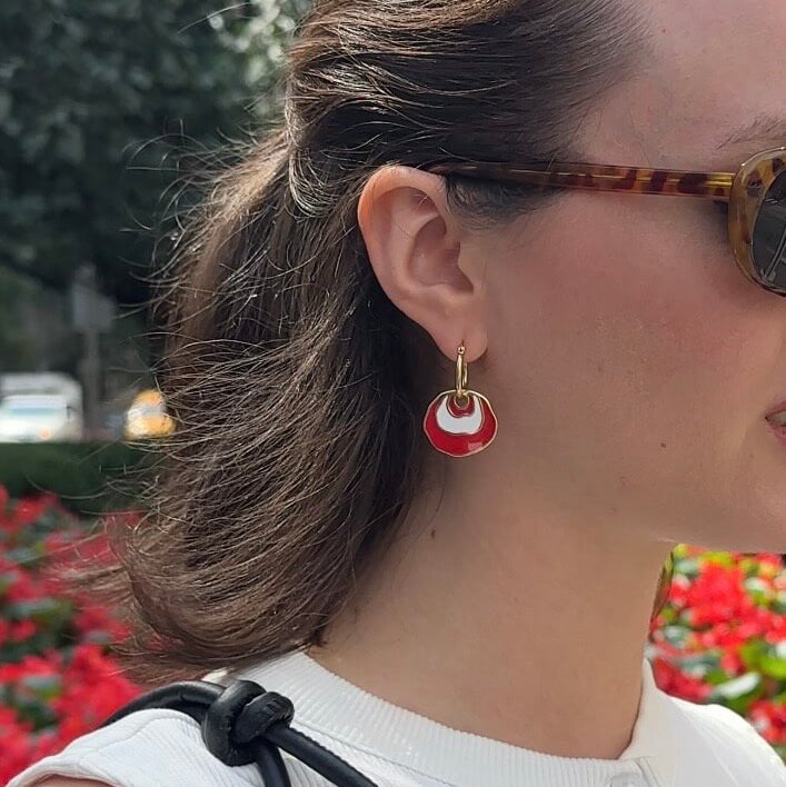 A close-up side profile of a woman outdoors wearing a gold hoop earring with a white and red concentric dangle design, along with tortoiseshell sunglasses and a white top.