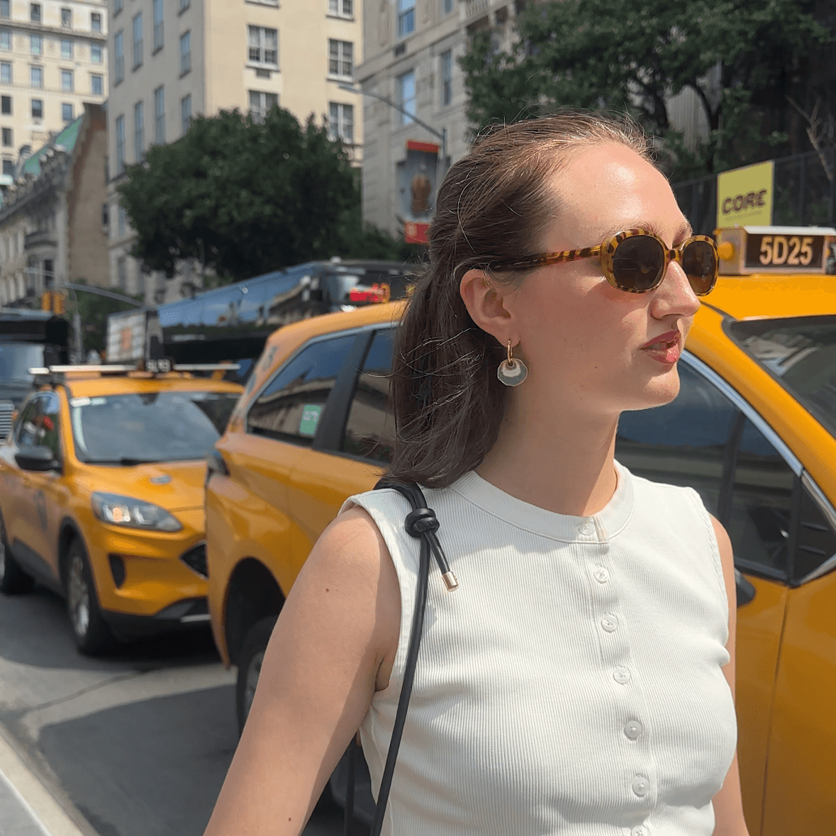 A woman wearing a white sleeveless button-front top and a gray pleated skirt walks on a city street with yellow taxis and tall buildings in the background. She is wearing a gold hoop earring with a white and light green concentric enamel dangle design.