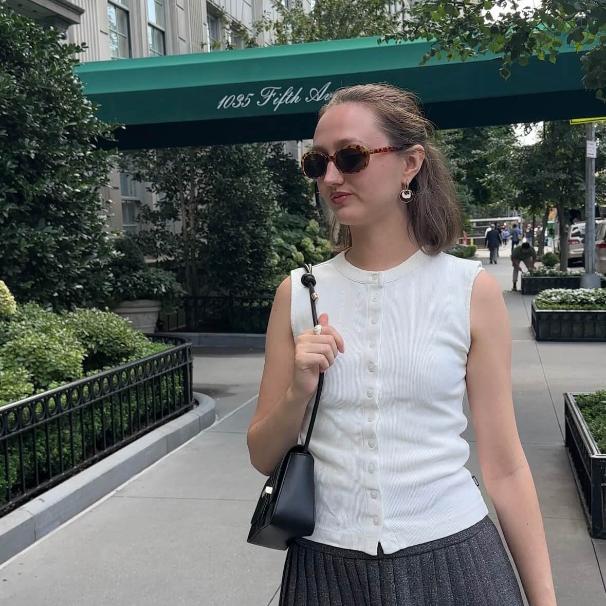 A woman wearing a white sleeveless button-front top and a dark gray pleated mini skirt, standing on a city sidewalk. She is wearing sunglasses and carrying a small black shoulder bag. She is wearing a brown and white enamel earring with a wavy circular shape.