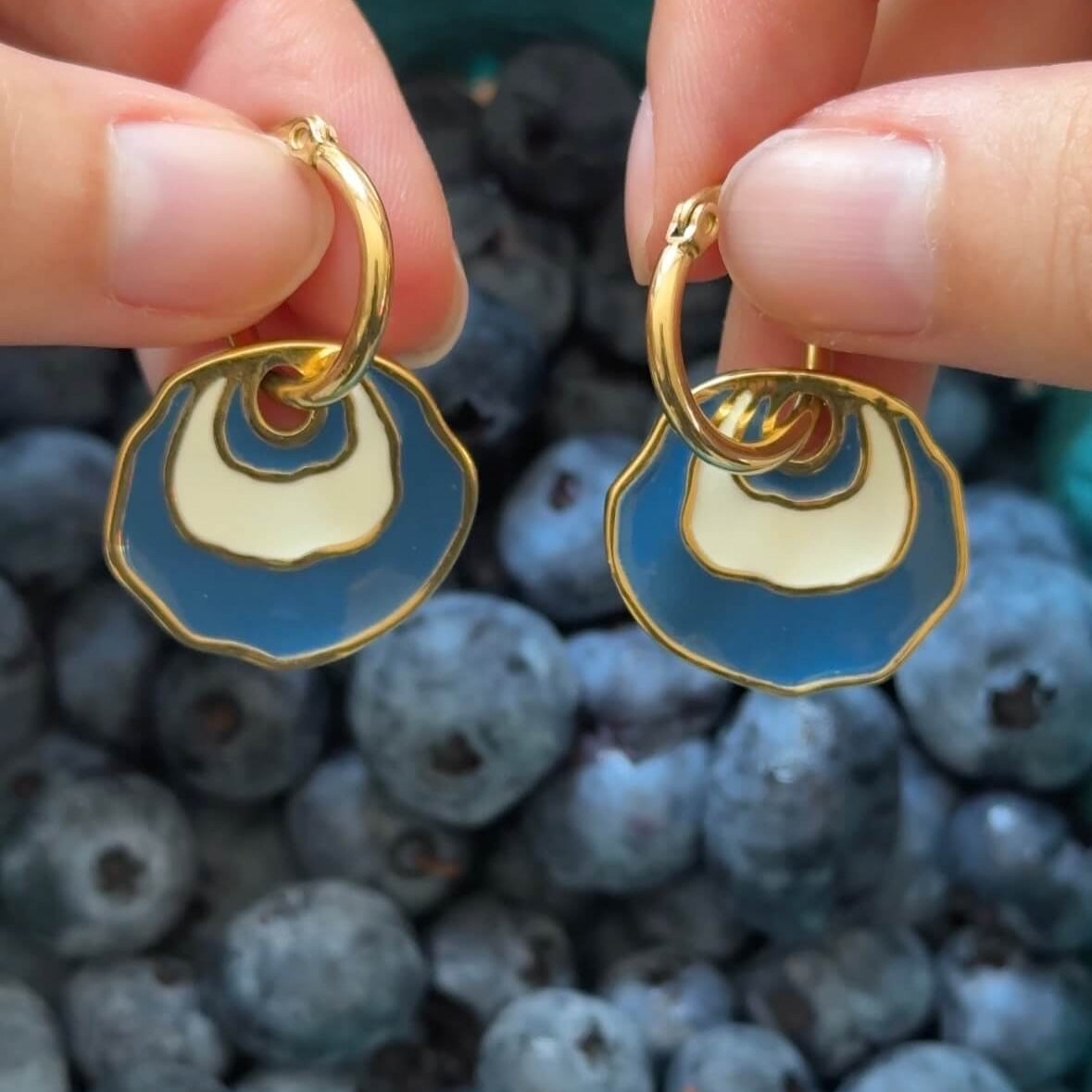 A close-up of a person's hands holding a pair of gold hoop earrings with a white and blue concentric enamel dangle design in front of a pile of blueberries.