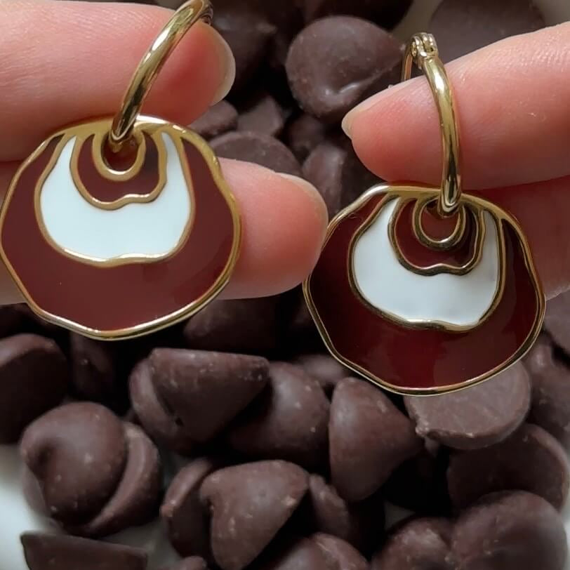 A pair of dangle earrings held between fingers, featuring a brown and white concentric circle design with gold metal borders and hoops, set against a background of chocolate chips.