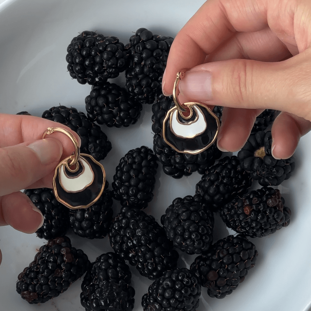 Gold hoop earrings with a white and black concentric enamel dangle design are held by hands over a white bowl filled with blackberries on a white bowl.