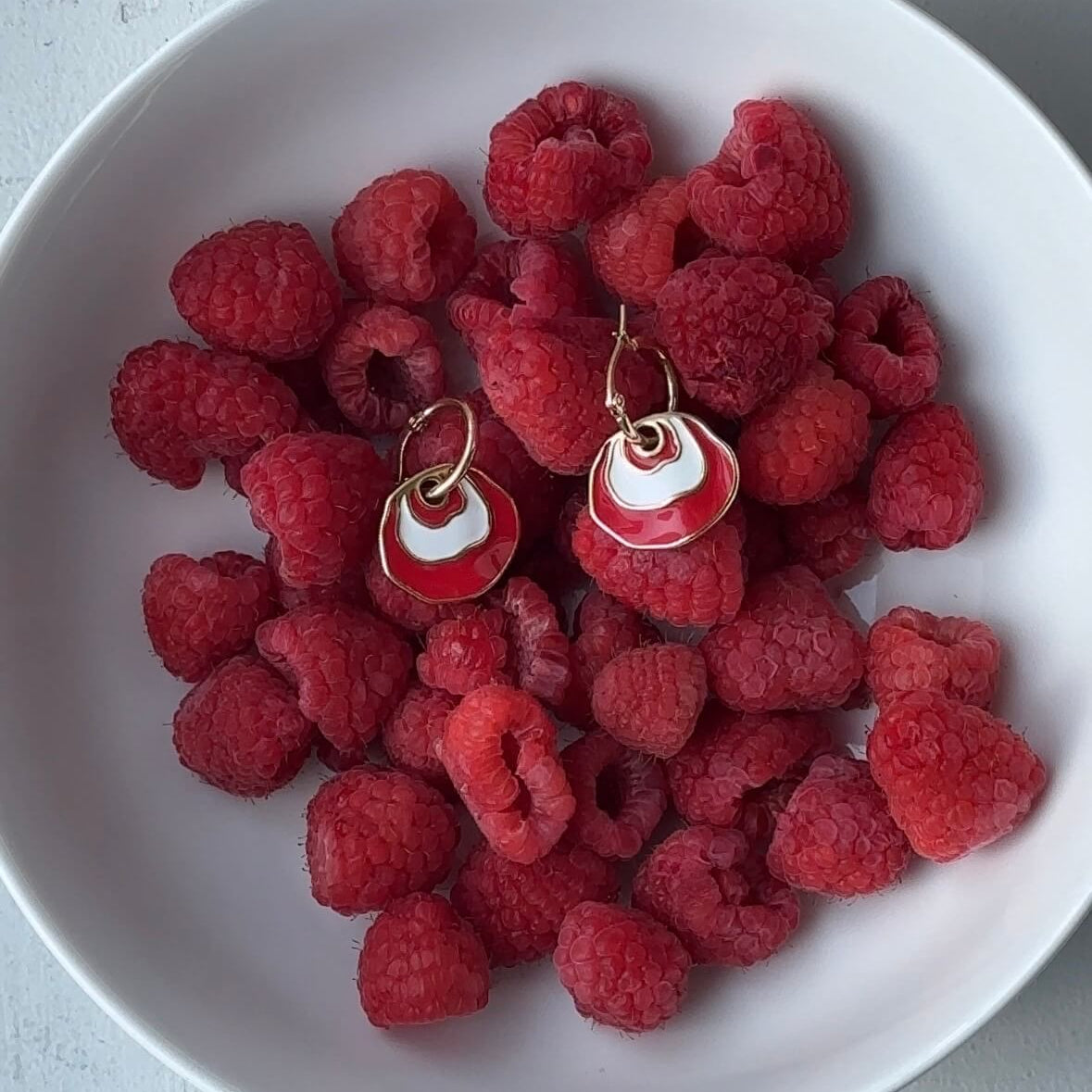 Gold hoop earring with a white and red concentric dangle design resting on a pile of fresh red raspberries in a white bowl.