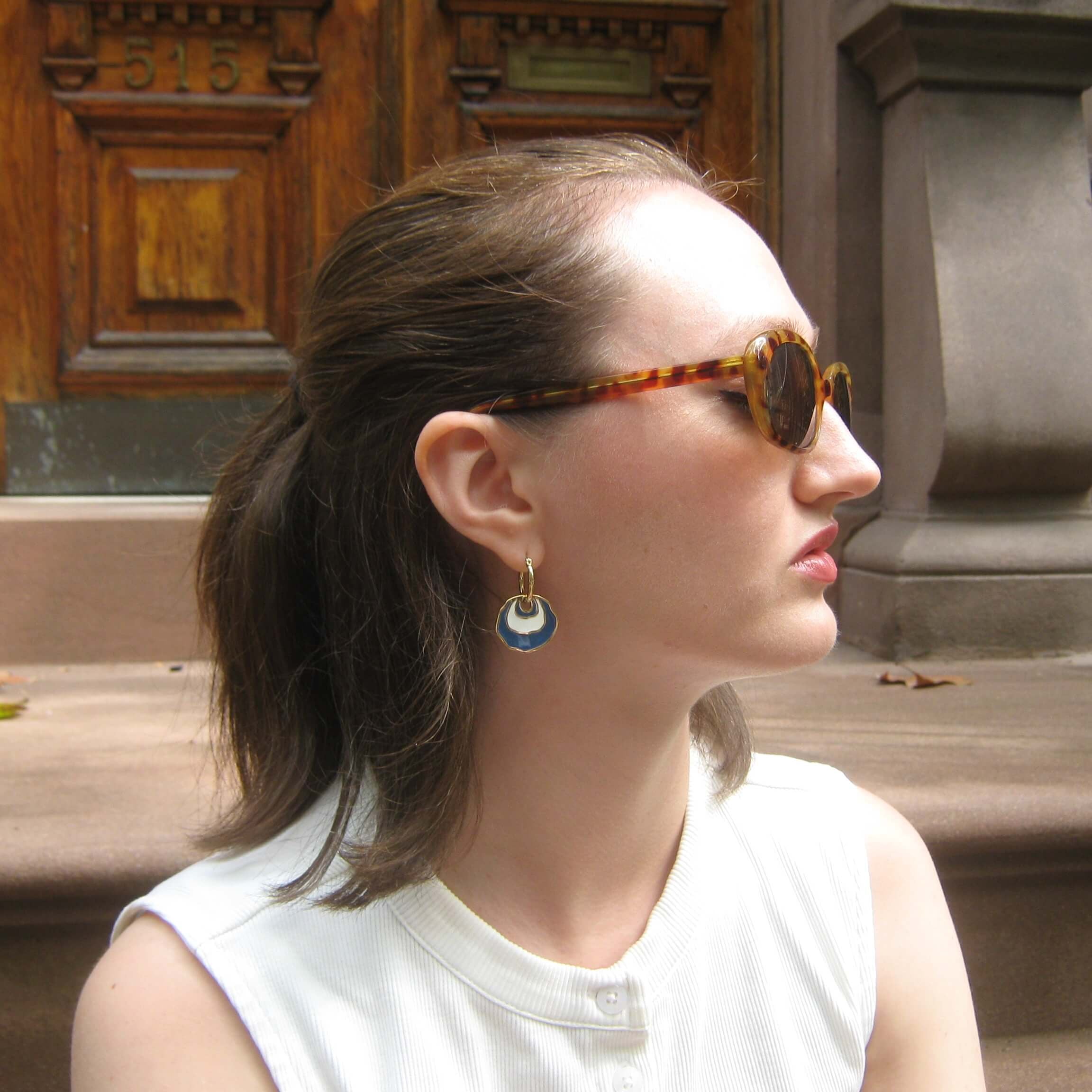 A woman in profile wearing tortoiseshell sunglasses and gold hoop earrings with a white and blue concentric enamel dangle design, sitting outdoors in front of a brownstone building.