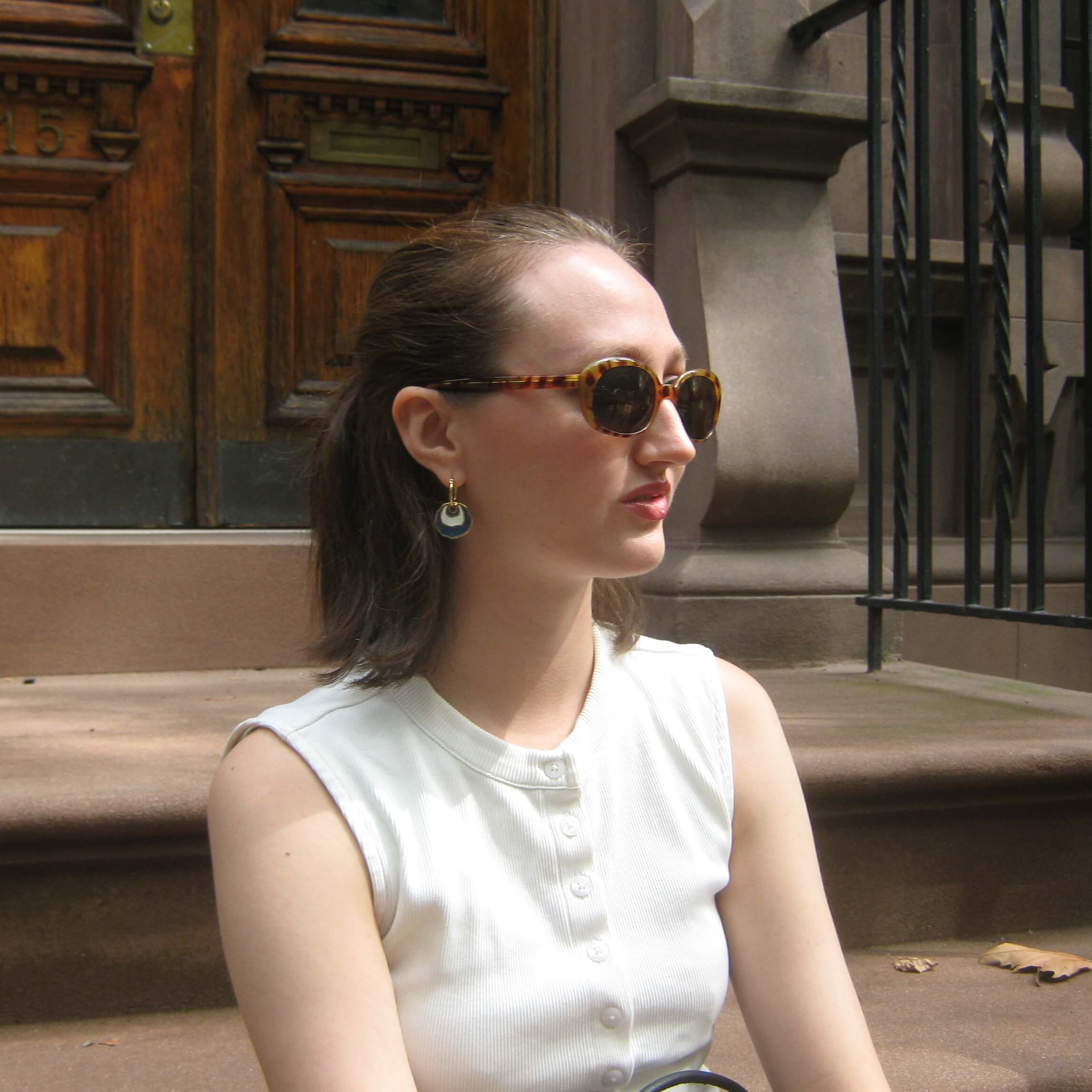 A woman with brown hair in a ponytail wearing tortoiseshell sunglasses and a white sleeveless top, sitting on stone steps in front of a brown wooden door. She is wearing a gold hoop earring with a white and blue concentric enamel dangle design.