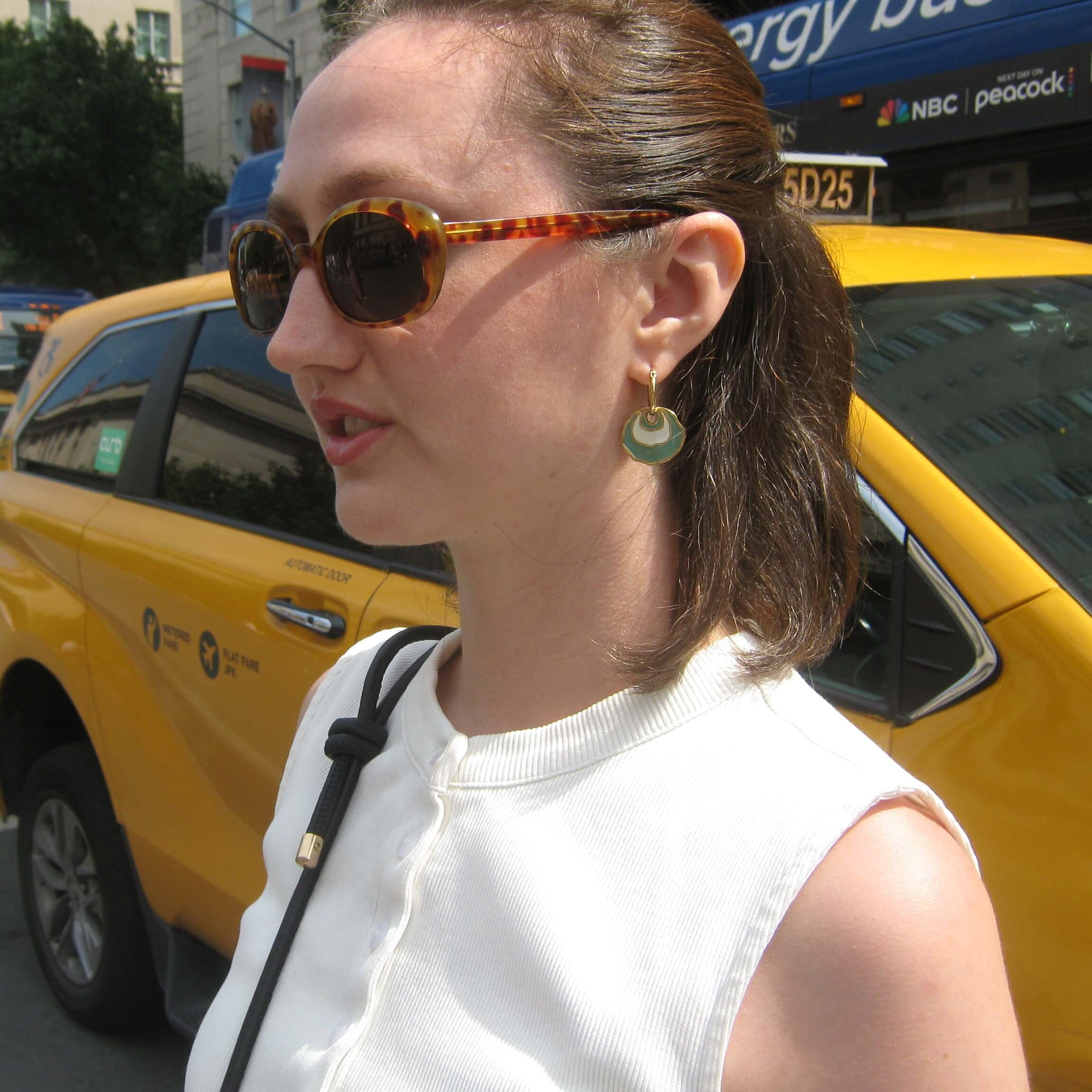 A woman wearing tortoiseshell sunglasses and a white sleeveless top stands on a city street with yellow taxis in the background. She is wearing a gold hoop earring with a white and light green concentric enamel dangle design.