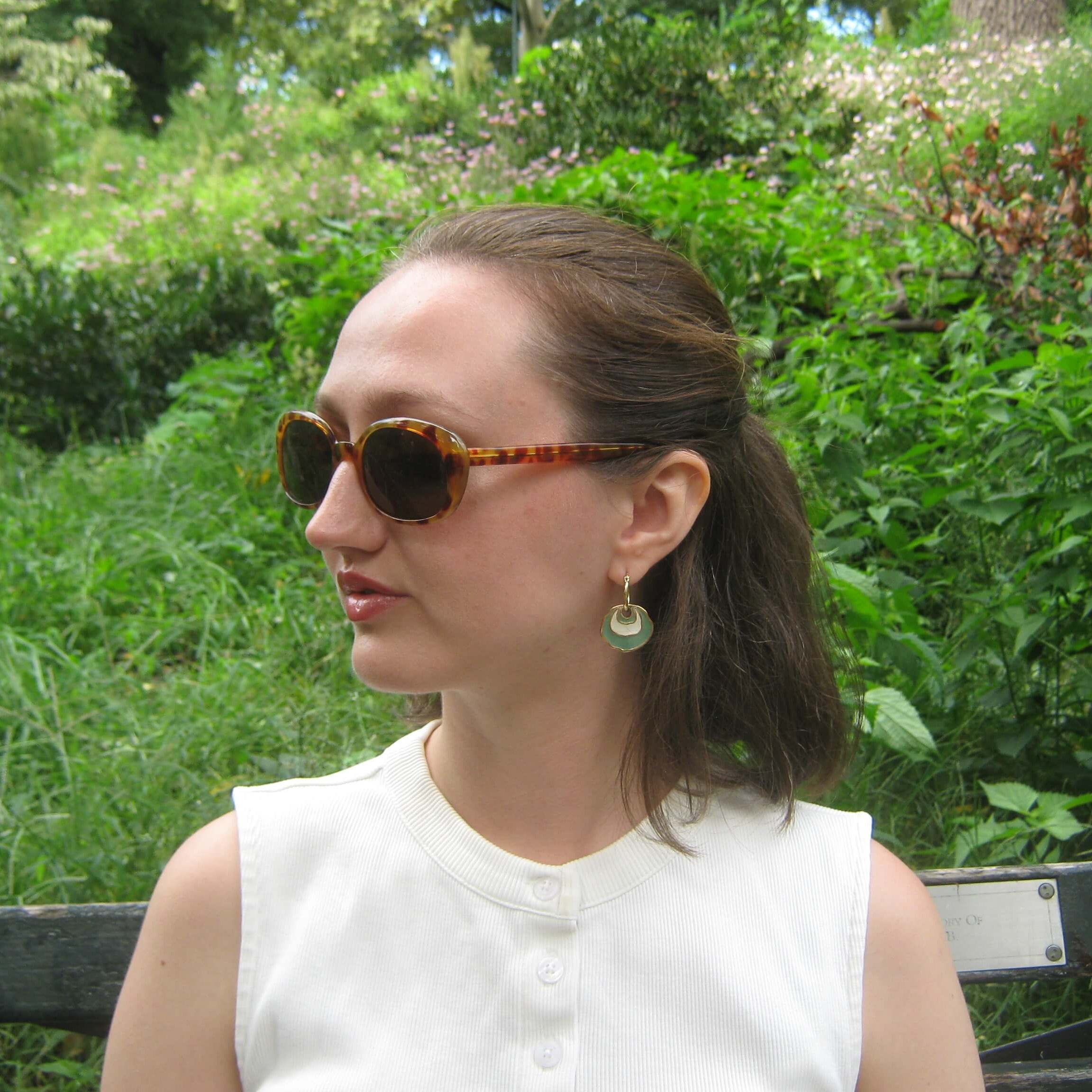 A woman wearing tortoiseshell sunglasses and a white sleeveless top, sitting on a bench in a lush green outdoor garden setting. She is wearing a gold hoop earring with a white and light green concentric enamel dangle design.