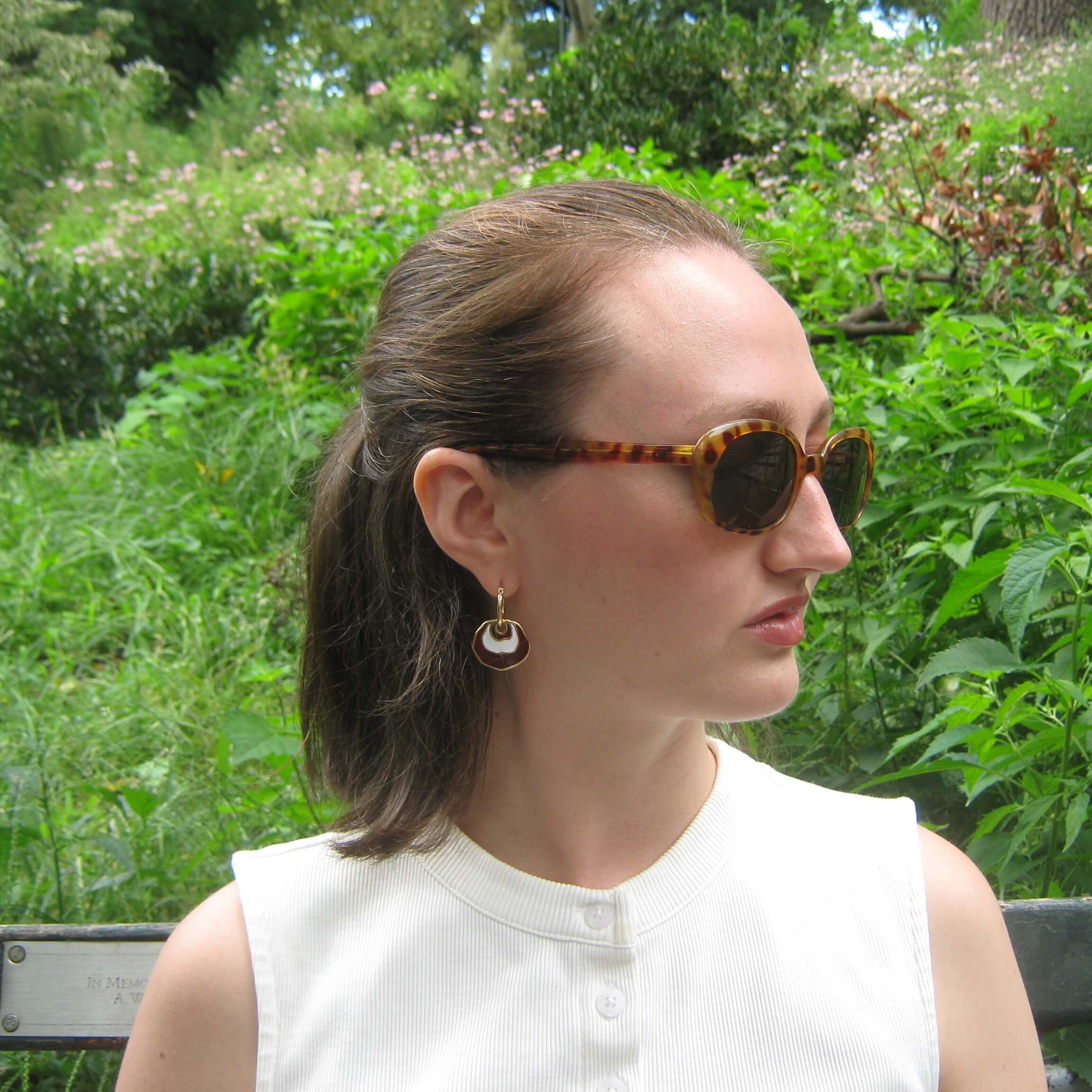 A woman in a white sleeveless top wearing tortoiseshell sunglasses and a hoop earrings with a brown and white enamel concentric dangle design, photographed outdoors in a lush green garden setting.