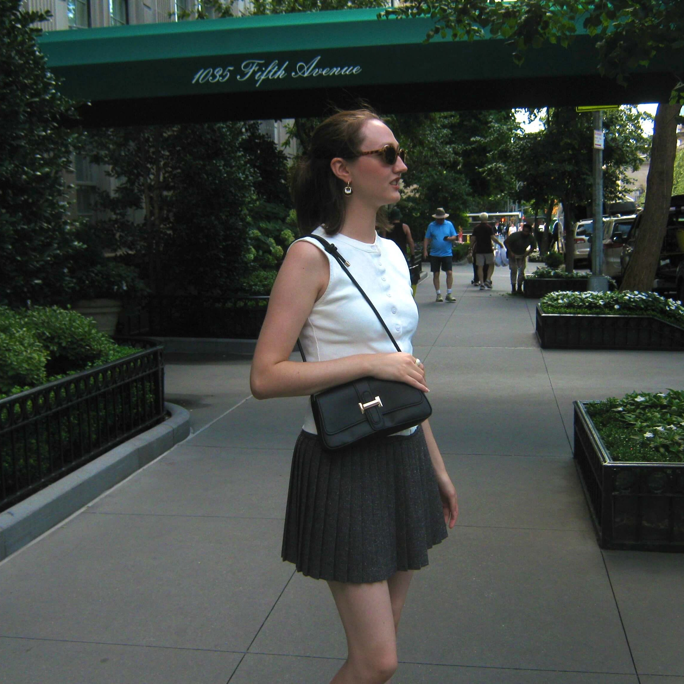 A woman wearing a white sleeveless top and a dark pleated mini skirt walks on a city sidewalk, carrying a black shoulder bag with a gold clasp. She is pictured outdoors under a green awning with '1035 Fifth Avenue' written on it. She is wearing a gold hoop earring with a white and brown concentric dangle design.